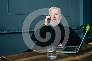 Tired gray-haired mature aged business man talking on mobile phone sitting at wooden table with laptop computer at home