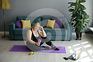 A tired happy fat woman is sitting on a training mat holding a bottle of water in her hand.