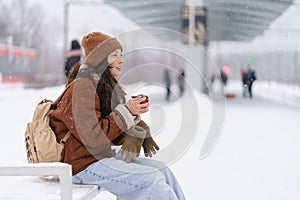 Tired but happy female traveler with mug of hot tea waiting for train to arrive in wintertime