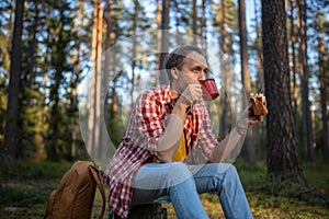 Tired hiker camper man eating sandwich drinking tea on break halt in forest enjoying nature.