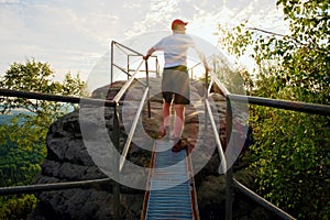 Tired hiker keep handrail on peak. Sunny spring daybreak in rocky mountains. Hiker with red baseball cap, dark pants and white