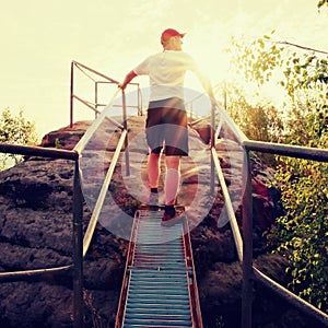 Tired hiker keep handrail on peak. Sunny spring daybreak in rocky mountains. Hiker with red baseball cap, dark pants and white shi