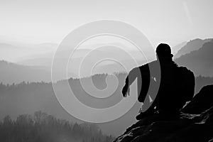 Tired hiker with sporty backpack sit on rocky peak and watching into deep misty valley bellow. Sunny spring daybreak in rocky