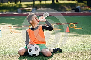 Tired hispanic boy in soccer uniform drinks water from plastic bottle after intensive training at stadium in morning.