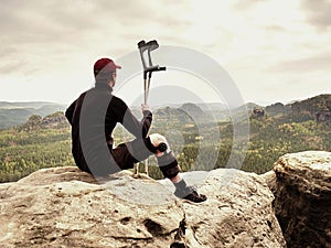 Tired hurt tourist with medicine crutches. Man with broken leg in knee brace features resting on exposed rocky summit