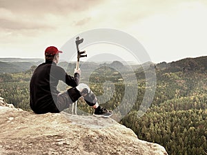 Tired hurt tourist with medicine crutches. Man with broken leg in knee brace features resting on exposed rocky summit