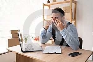 tired indian male small business owner working at laptop, online store delivery worker has vision problems, businessman