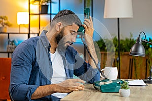 Tired Indian man freelancer talking on retro wired telephone annoyed boring conversation at office