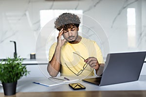 Tired indian man holding phone working on laptop computer at the table at home, drinking coffee at the kitchen