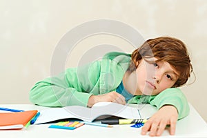 Tired kid sitting at the desk with many books. Sad schoolboy doing homework