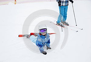 Tired kid in warm blue overalls fell and lies on the snow, resting from skiing lesson