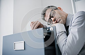 Tired lazy office worker leaning on a filing cabinet and sleeping, he is falling asleep standing up; stress, unproductivity and