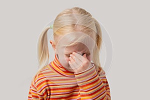 Tired little blonde girl covered her nose with her hand. Isolated on a gray background
