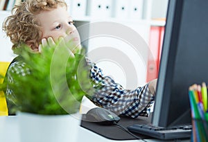 Tired little boy propped his head in his hands and looks at the monitor at the office