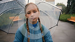 Tired little girl schoolgirl child looking at camera bored unhappy bad weather rain umbrella city park outdoors fatigue