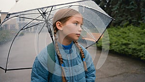 Tired little girl schoolgirl child looking at camera bored unhappy bad weather rain umbrella city park outdoors fatigue