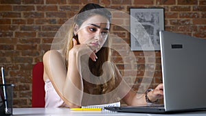 Tired look of caucasian girl who is working on her laptop and making pauses to relax while being in red brick studio