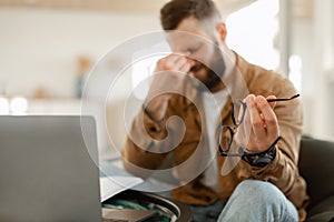 Tired Male Holding Glasses Massaging Nosebridge Sitting At Laptop Indoors