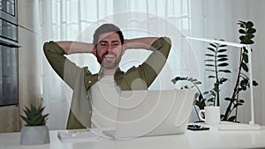 Tired male sitting at white desk while holding hands behind head at home office