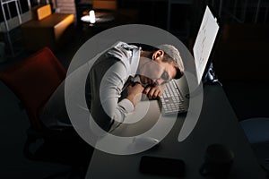 Tired male trader asleep on computer desk late at night alone. Exhausted yougng businessman sleeping with head on