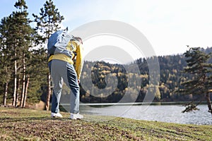 Tired male traveler with heavy backpack bending over near forest lake back view