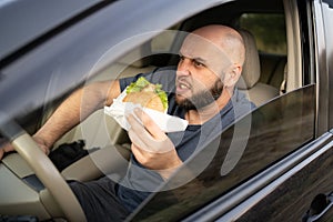 Tired man driving car while eating hamburger. Angry in waiting standing in traffic jam