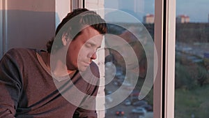 Tired man looks out the window sitting on the balcony at sunset in the evening.