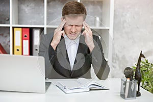 Tired man manager with laptop suffering terrible headache at his working place in office with his hands near his head looking