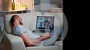 Tired man in pajamas sitting on sofa falling asleep while having online business videocall with teammates