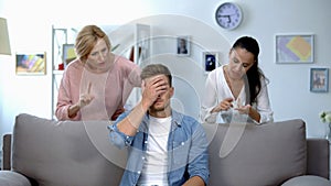 Tired man sitting on sofa, enduring to wife and mother criticize him, quarrel