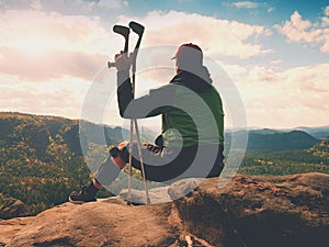 Tired man tourist sit with hurt knee in immobilizer or rock, hold medicine pole. Open forest landscape