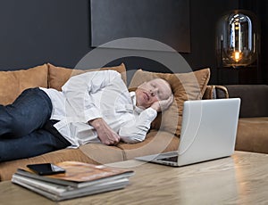 Tired mature businessman sleeping on couch in office or at home. Overtime work or overload at work concept.