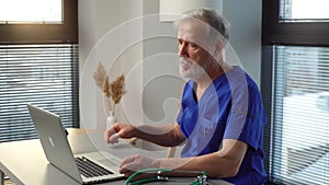 Tired mature male doctor sitting down at table, starting working typing on laptop computer.