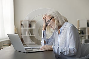 Tired mature senior business woman touching head at laptop