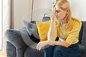 Tired mature woman sitting on the sofa at home, rubbing temple with suffering face, feels sick and weary, senior female