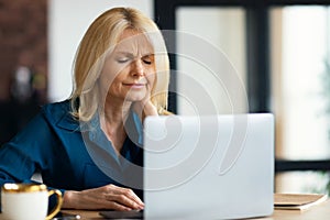Tired mature woman suffering from neck spasm while working on laptop at home office, sitting at desk and rubbing muscles