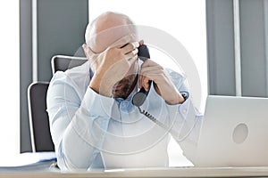 Tired mid adult businessman using landline phone at desk in office