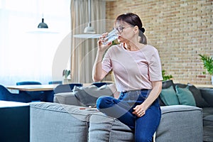 Tired middle aged woman drinking water from glass, sitting at home