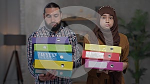 Tired Middle Eastern couple standing in home office holding document folders sighing. Portrait of overwhelmed man and