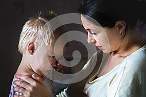 Tired mother hugging and calming little cute sad upset crying caucasian blond toddler boy at home indoor. Mom holding sleepy sick