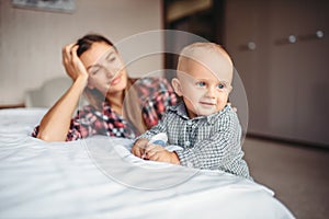 Tired mother looks on her child playing on the bed