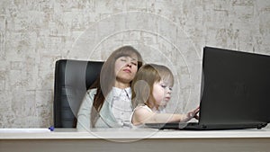 Tired mother works at the table with her little daughter on the computer in the office. business woman working on a