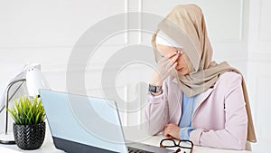 Tired muslim woman works and types on laptop, puts off her glasses and rubs her eyes.