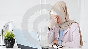 Tired muslim woman works and types on laptop, puts off her glasses and rubs her eyes.