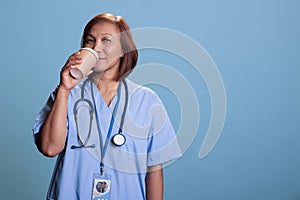 Tired nurse wearing blue uniform and stethoscope drinking coffee in front of camera