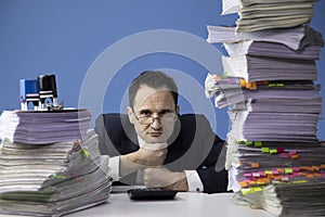 Tired office worker puts his head on his fists, amidst high stacks of documents