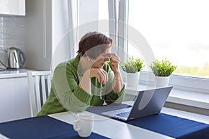 Tired old woman in front of a monitor is massaging the bridge of her nose.
