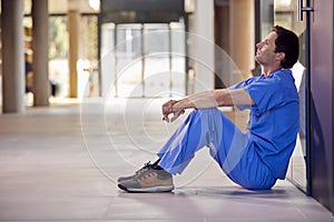 Tired And Overworked Medical Worker In Scrubs Sitting On Floor Of Hospital Corridor During Break