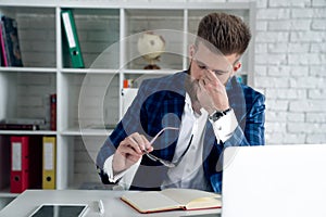 Tired and overworked stylish red bearded manager sitting in front of computer and trying to concentrate, his eyes are dry and hurt