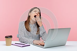 Tired overworked woman office worker yawning sitting at workplace with laptop, exhausted with job routine, showing disinterest